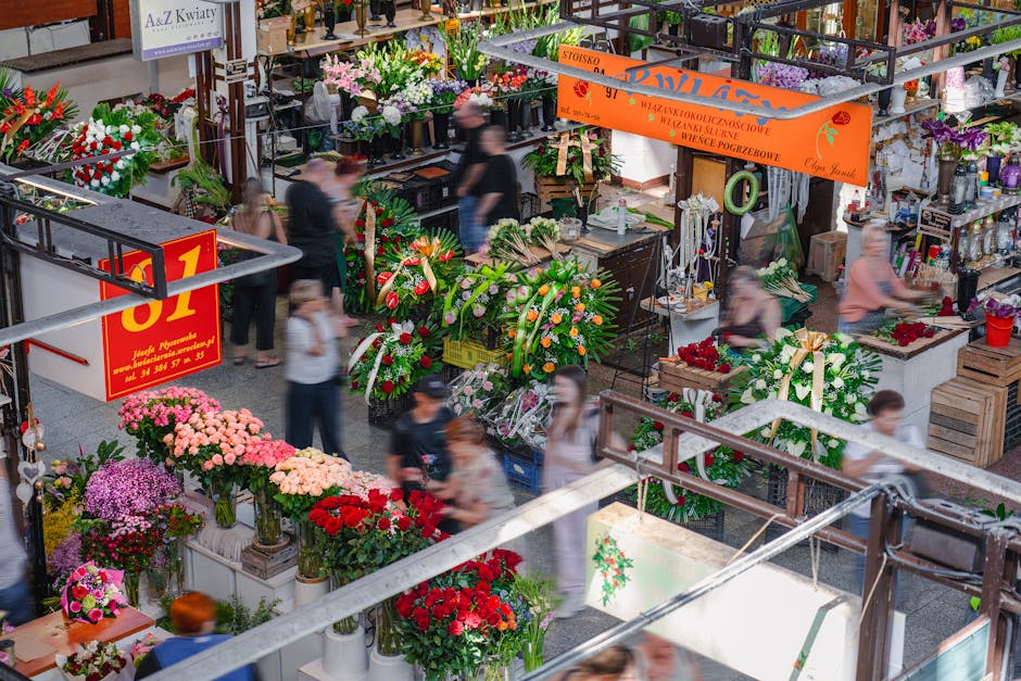 A busy indoor flower market scene featuring a variety of fresh floral arrangements and bouquets displayed on tables and shelves. Prominent flowers include pink chrysanthemums, red roses, purple chrysa
